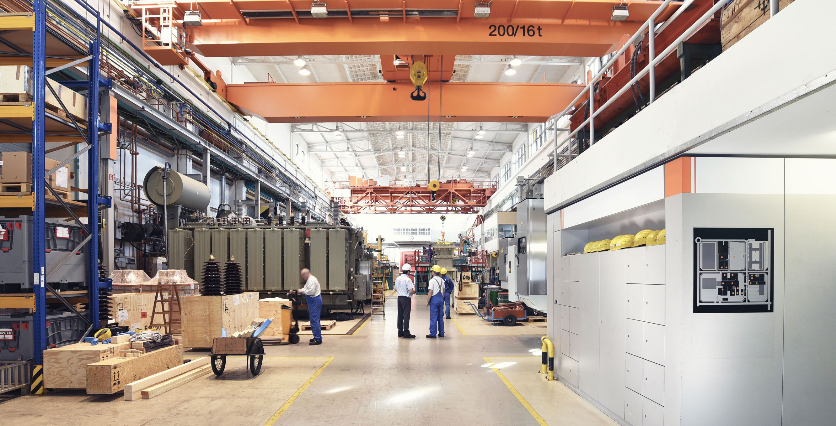 A spacious factory hall with people working at machines. A crane bears the inscription "200/16t". Shelving units and machines are visible on both sides, with plenty of light coming from above.