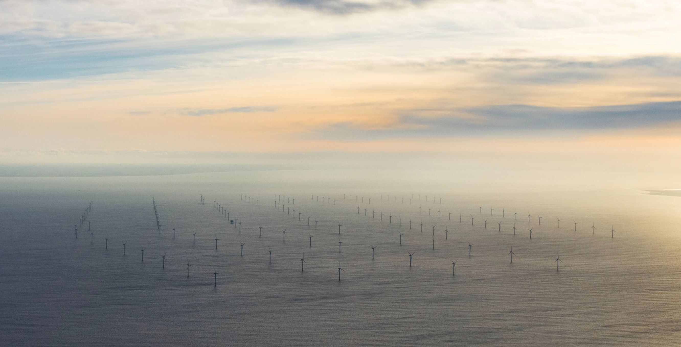 Ein Luftbild zeigt einen großen Offshore-Windpark mit zahlreichen Windturbinen, die im Meer stehen. Der Himmel ist teilweise bewölkt, und die Sonne scheint durch, was eine sanfte Beleuchtung erzeugt.