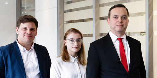 Drei Personen stehen vor einer Glaswand. Links steht eine Person in einem blauen Anzug, in der Mitte eine Person mit Brille und weißem Hemd, und rechts eine Person in einem schwarzen Anzug mit roter Krawatte.