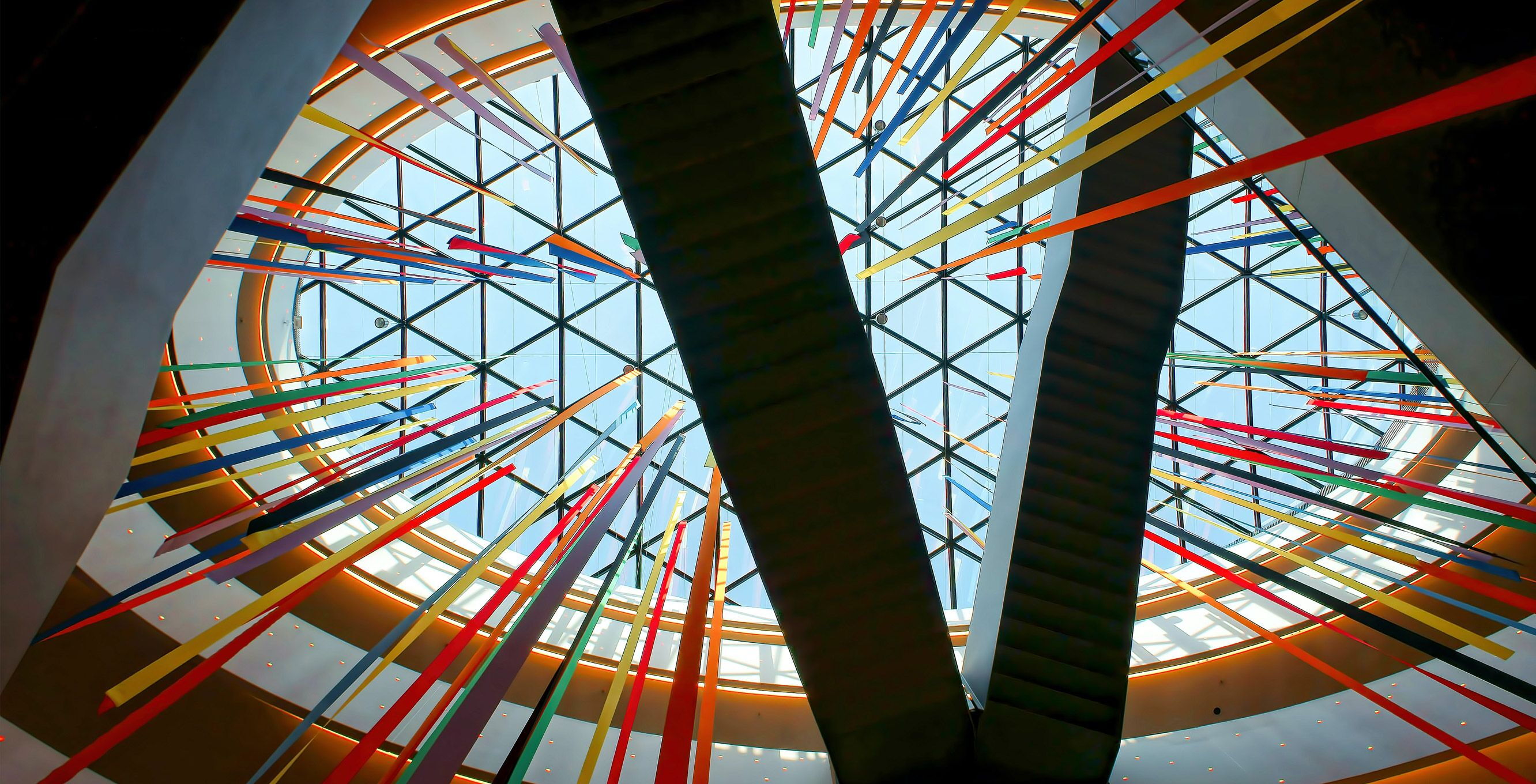 Blick nach oben durch ein gläsernes Dach, durch das bunte Bänder hängen. Zwei breite, dunkle Treppen kreuzen sich im Vordergrund und führen nach oben. Das Licht strömt durch das Glas und beleuchtet die Szenerie.