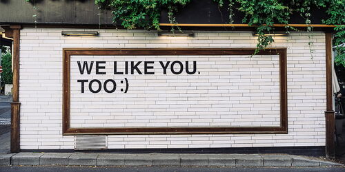 Weiße Ziegelwand mit einem Holzrahmen, darauf steht in schwarzen Großbuchstaben: "WE LIKE YOU, TOO ;)". Oben wachsen Pflanzen über den Rand der Wand. Darunter ist ein schmaler Bürgersteig zu sehen.