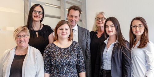A group of seven people—four women in the front and three people, including two women and a man, in the back—is standing in an office in front of a wall. Everyone is smiling at the camera.