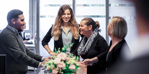 Fünf Personen stehen lächelnd um einen Schreibtisch in einem Büro. Im Vordergrund ist ein Blumenstrauß. Große Fenster im Hintergrund sorgen für viel Tageslicht. Die Gruppe wirkt freundlich und in ein Gespräch vertieft.