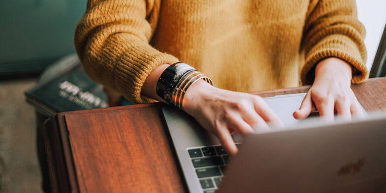 Eine Person in einem senfgelben Pullover sitzt an einem Holztisch und tippt auf einem Laptop. Ihr Arm ist mit mehreren Armbändern geschmückt. Der Hintergrund ist unscharf.