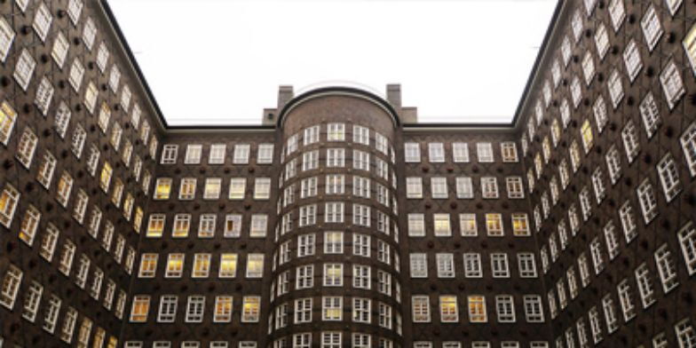 Backsteinfassade eines Gebäudes mit vielen Fenstern. Die Architektur zeigt eine symmetrische, gewölbte Form in der Mitte und zwei Flügel, die nach außen ragen. Einige Fenster sind beleuchtet. Der Himmel ist bedeckt.