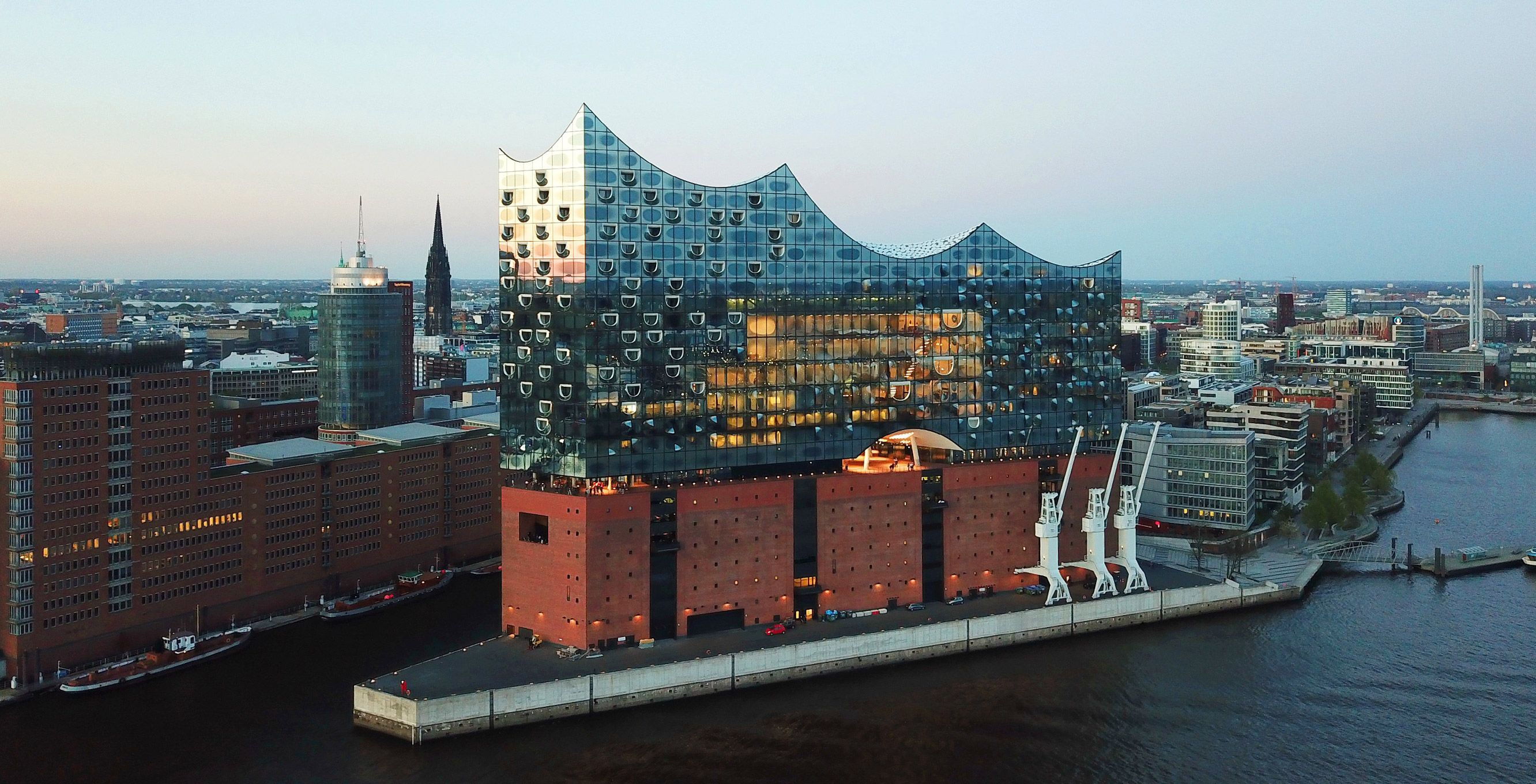 Luftaufnahme eines modernen, geschwungenen Gebäudes mit gläserner Fassade auf einem alten Backsteinsockel am Wasser, umgeben von Stadtarchitektur. Es ist Abend, und Licht spiegelt sich im Glas wider.