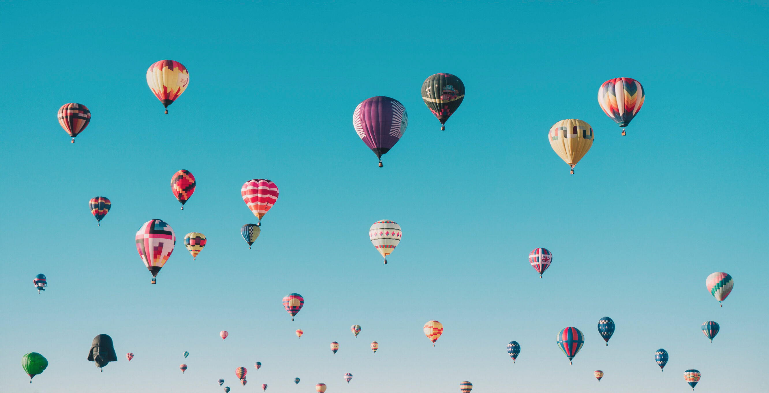 Mehrere bunte Heißluftballons in unterschiedlichen Designs schweben am Himmel vor einem klaren blauen Hintergrund.