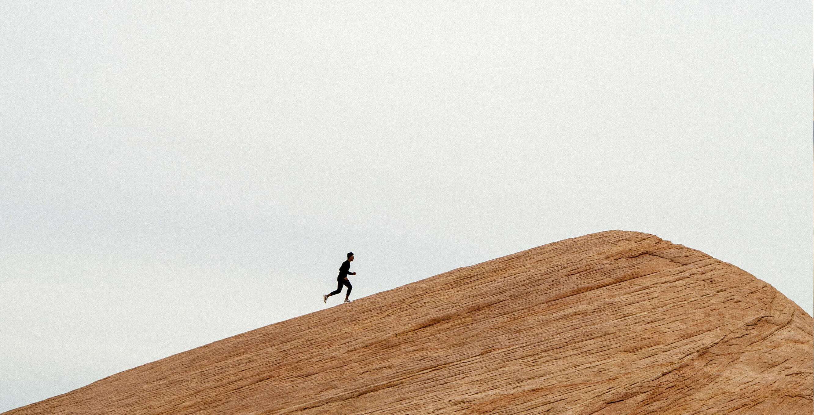 A person is walking up a rocky, sloping hill. The sky is overcast. The scene has a minimalist feel, and the person appears as a silhouette.