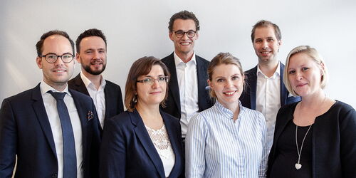 Eine Gruppe von sieben Personen in Business-Kleidung steht vor einer weißen Wand. Vier Männer und drei Frauen lächeln in die Kamera. Alle tragen formelle Kleidung, wie Anzüge und Blusen.
